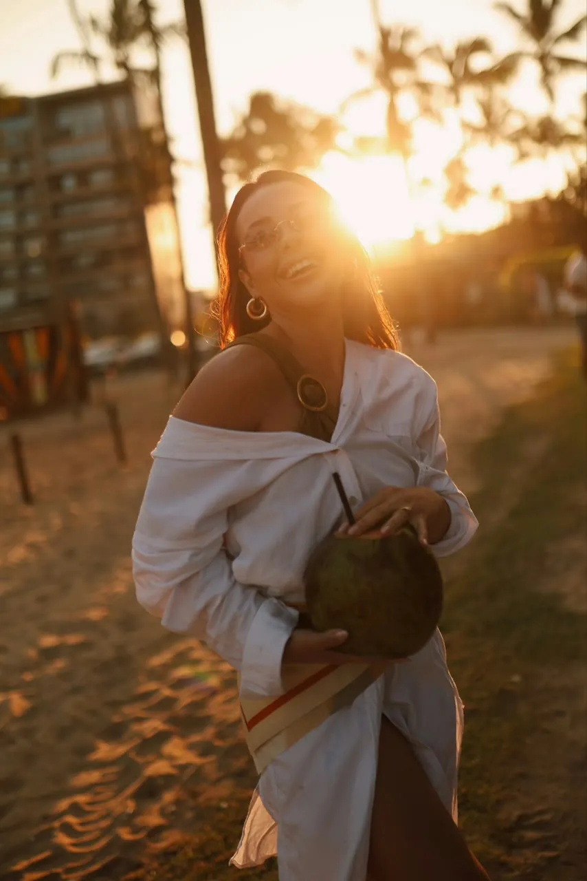Dra. Laisy Velar sorrindo com um coco na mão no pôr do sol