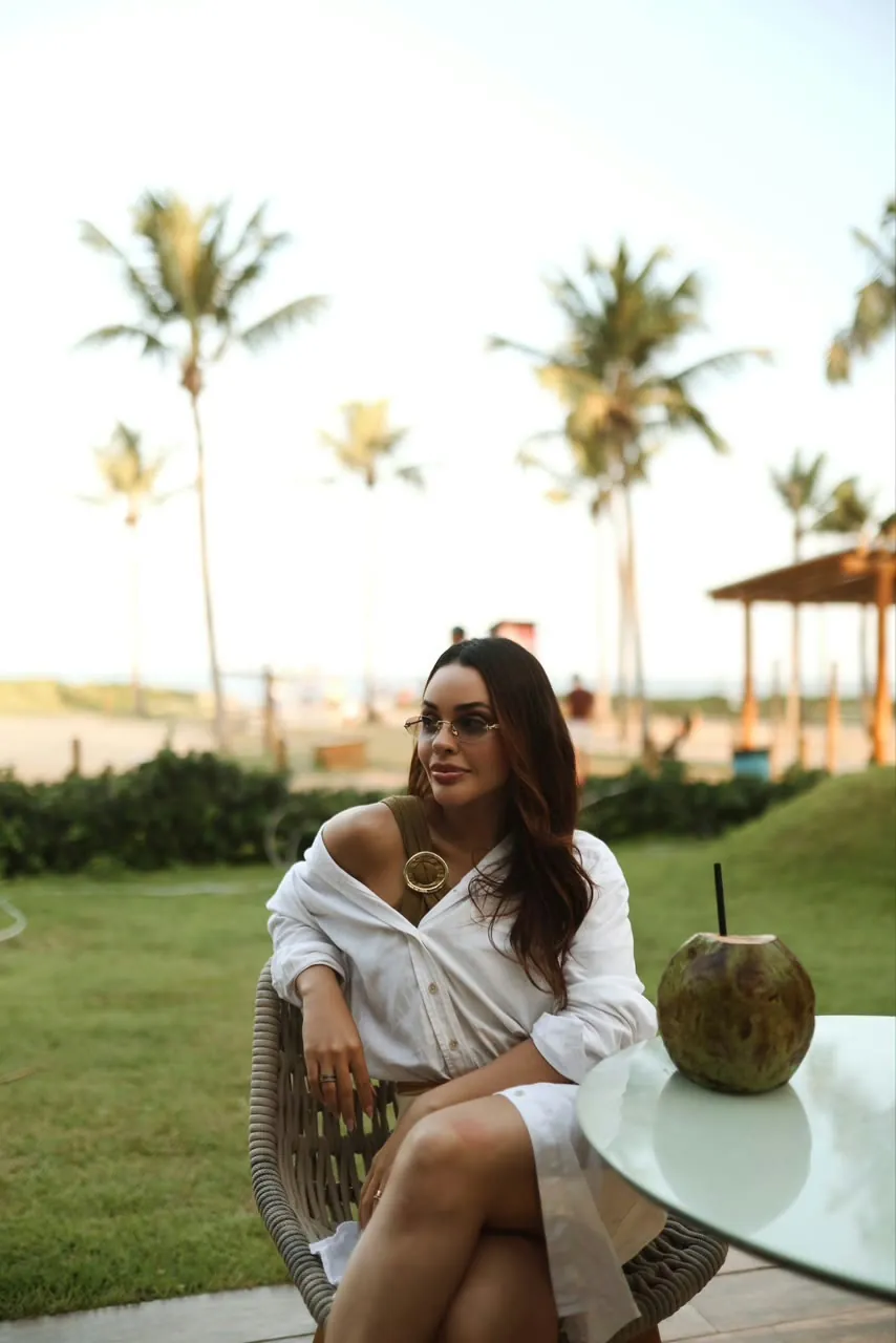 Dra. Laisy Velar sentada à beira-mar com um coco na mesa